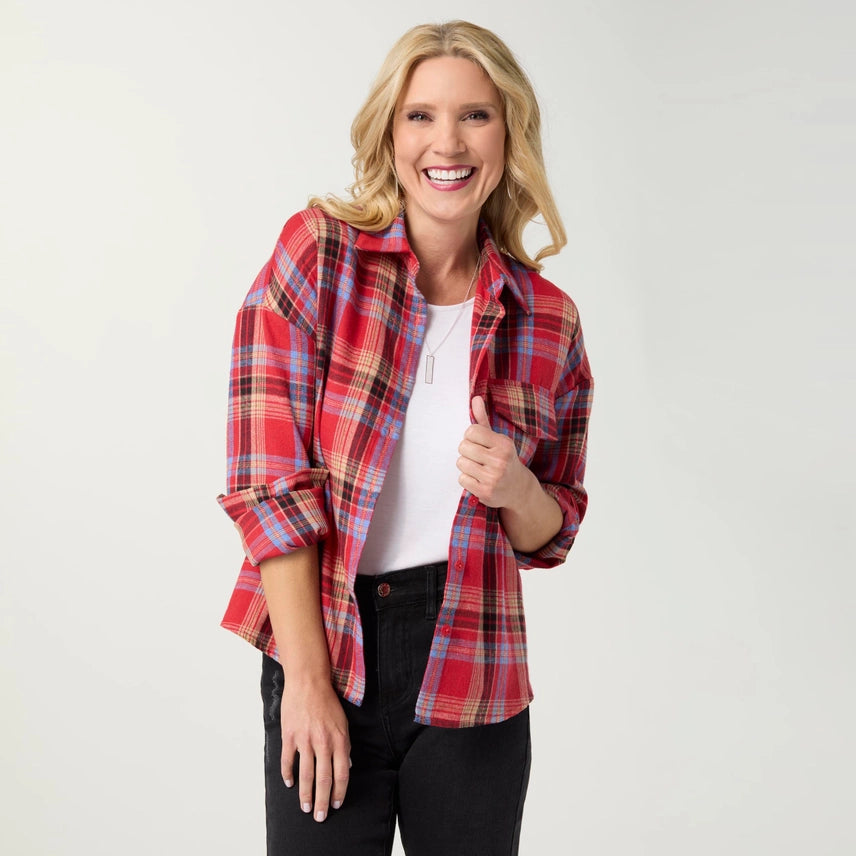 A smiling blonde woman wearing a red and blue plaid button-down shirt over a white t-shirt and black pants, standing against a light gray studio background.