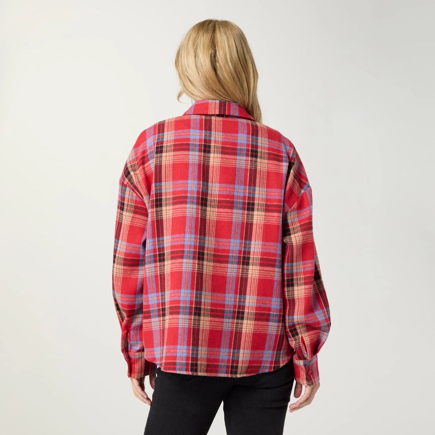 A woman viewed from behind wearing a red plaid button-up shirt with blue, beige, and black stripes, paired with black pants, against a plain light gray background.